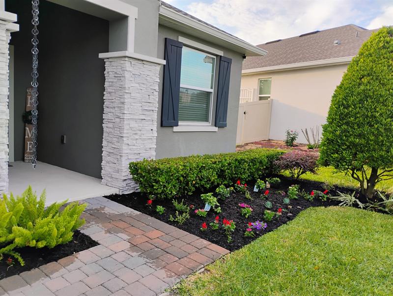 Dark mulch bed with red and purple flowers, trimmed shrubs and a topiary beside a stone-column porch